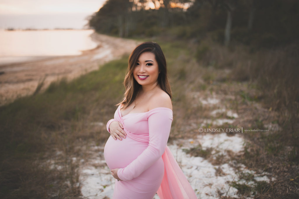 mama to be wearing a long flowey pink dress on a grassy shore