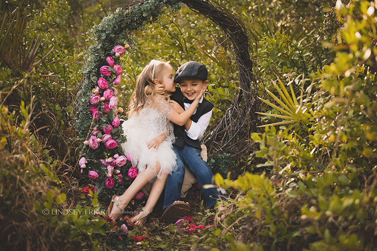 little girl kissing boy on cheek laughing on garden floral swing