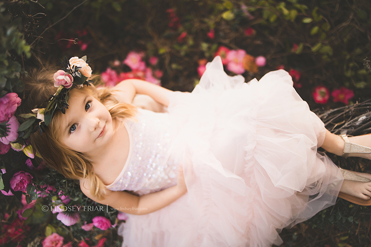 Dark floral background little girl with blonde hair and blue eyes wearing light pink sequin dress looking at camera