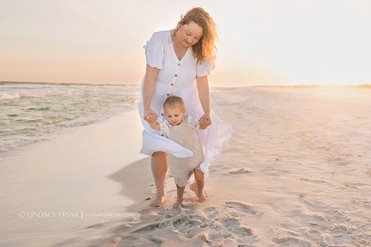 mother holding her baby's hands walking behind him on the beach